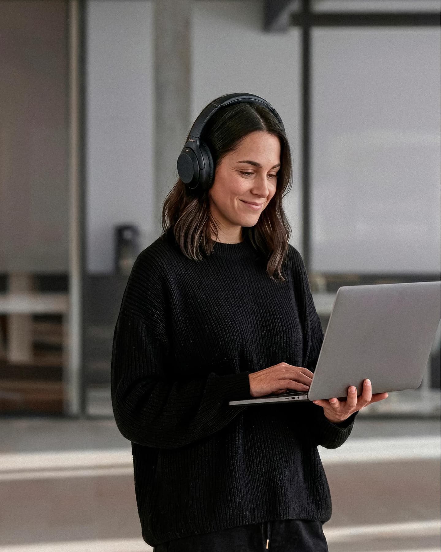Mujer con auriculares trabajando de pie con su notebook en el coworking, con mamparas vidriadas de fondo