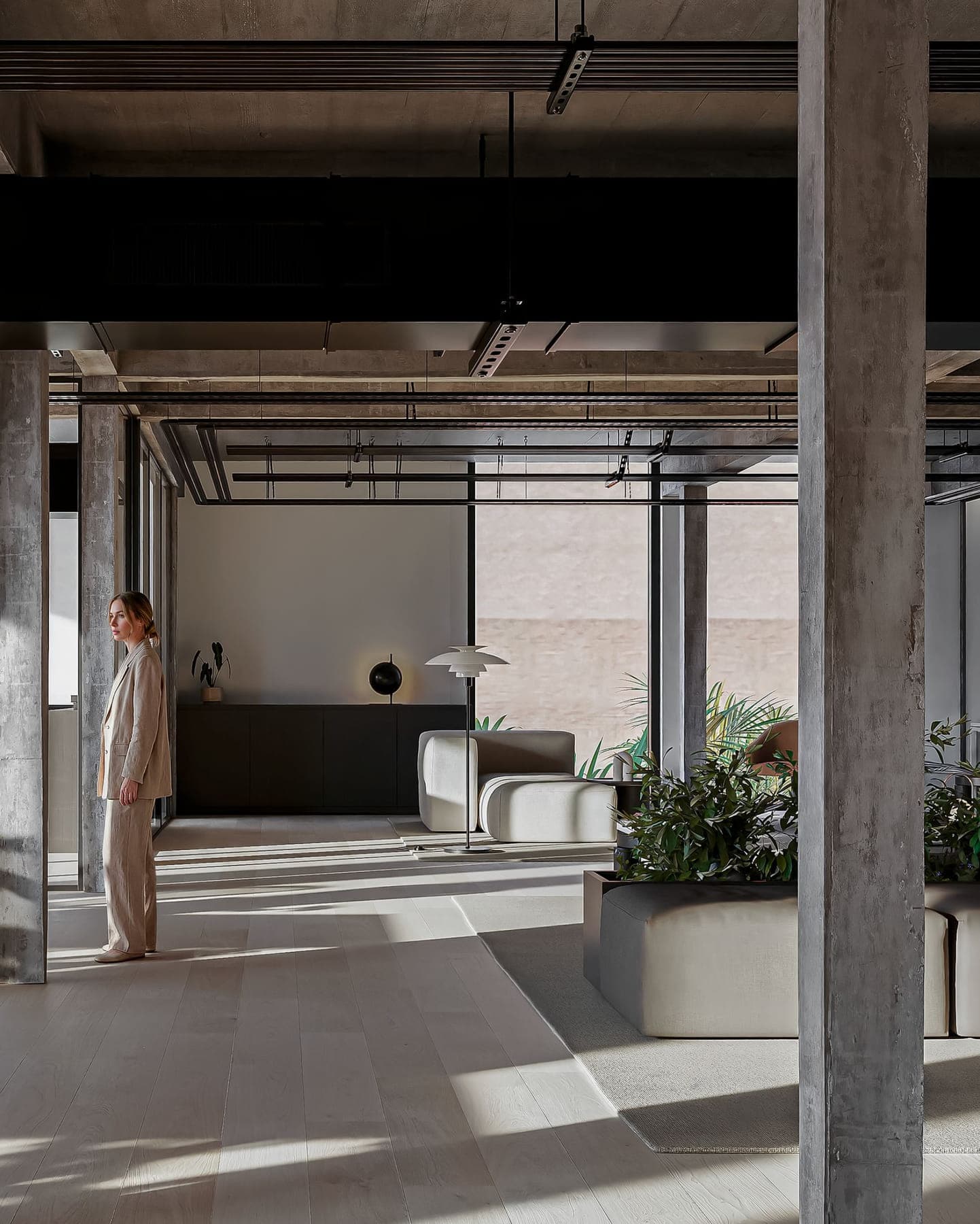 Ejecutiva de pie junto a los ventanales de una planta completa, con lounge de tapizados claros y luz de tarde